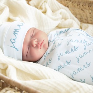 May include: A sleeping baby wrapped in a light blue swaddle blanket and matching hat. The name "James" is written in a script font on both the hat and blanket. The baby is nestled in a woven basket lined with a cream-colored fabric.