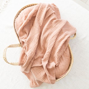 May include: A light pink muslin swaddle blanket in a woven wicker basket.