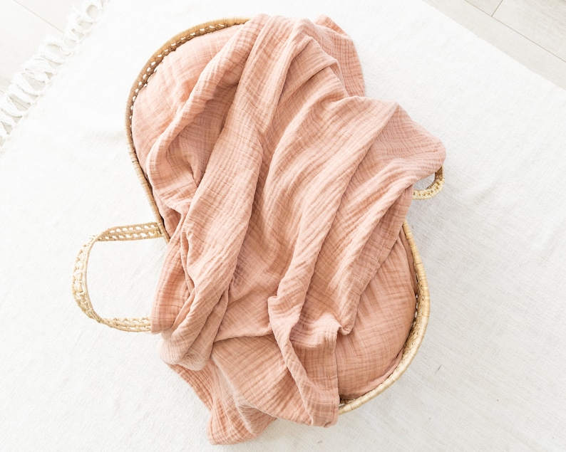 May include: A light pink muslin swaddle blanket in a woven wicker basket.