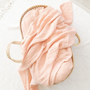 May include: A peach-coloured muslin swaddle blanket is folded and placed inside a woven basket with handles.