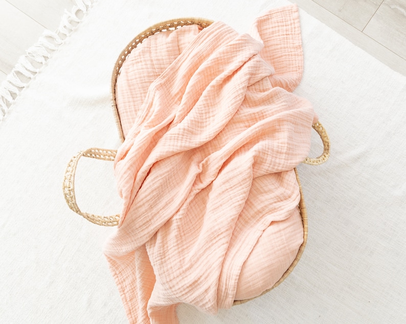 May include: A peach-coloured muslin swaddle blanket is folded and placed inside a woven basket with handles.
