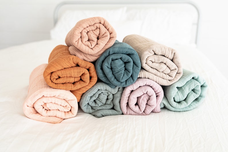May include: A stack of eight rolled up muslin blankets in various colours, including peach, orange, teal, and light pink. The blankets are arranged in a pyramid shape on a white bed.
