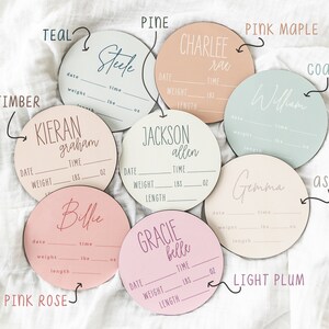 May include: Ten round wooden baby announcement plaques in various colors, including teal, pine, pink maple, coast, ash, light plum, and pink rose. Each plaque has a blank space for a name, date, time, weight, lbs, oz, and length.