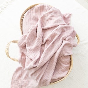 May include: A pink muslin swaddle blanket in a woven basket. The blanket is soft and textured, and the basket is made of natural materials.