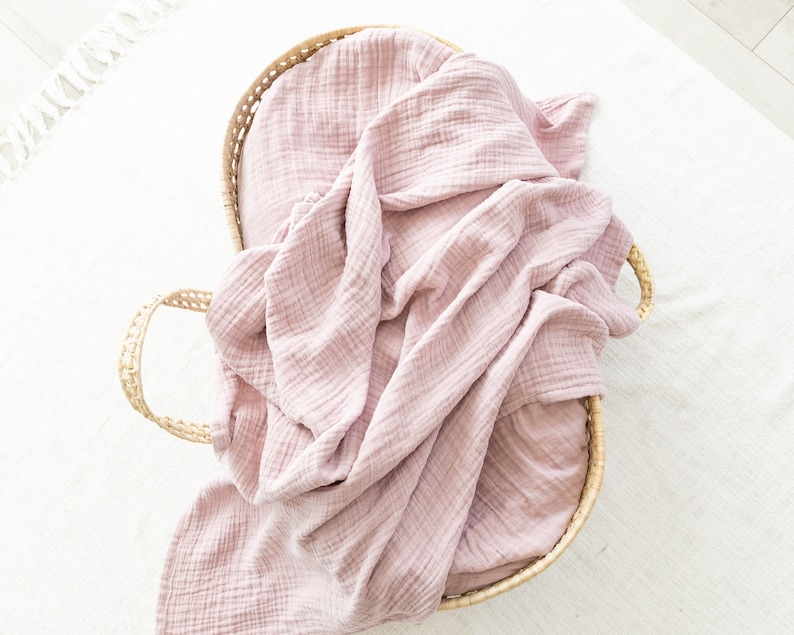 May include: A pink muslin swaddle blanket in a woven basket. The blanket is soft and textured, and the basket is made of natural materials.