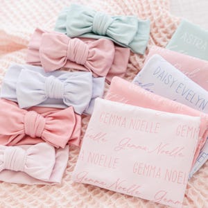 May include: A collection of baby headbands and blankets in pastel colors. The headbands are light pink, blue, and mint green with large bow details. The blankets are pink and blue, with names printed on them in a cursive font.