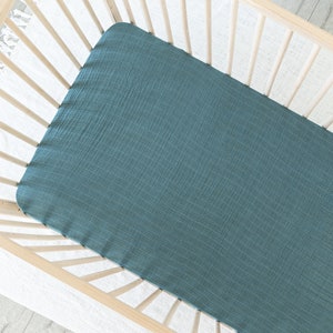 May include: A teal-colored fitted crib sheet made of a textured fabric. The sheet is placed inside a light-colored wooden crib, creating a soft and inviting look for a baby's nursery. The sheet's color adds a touch of calm.