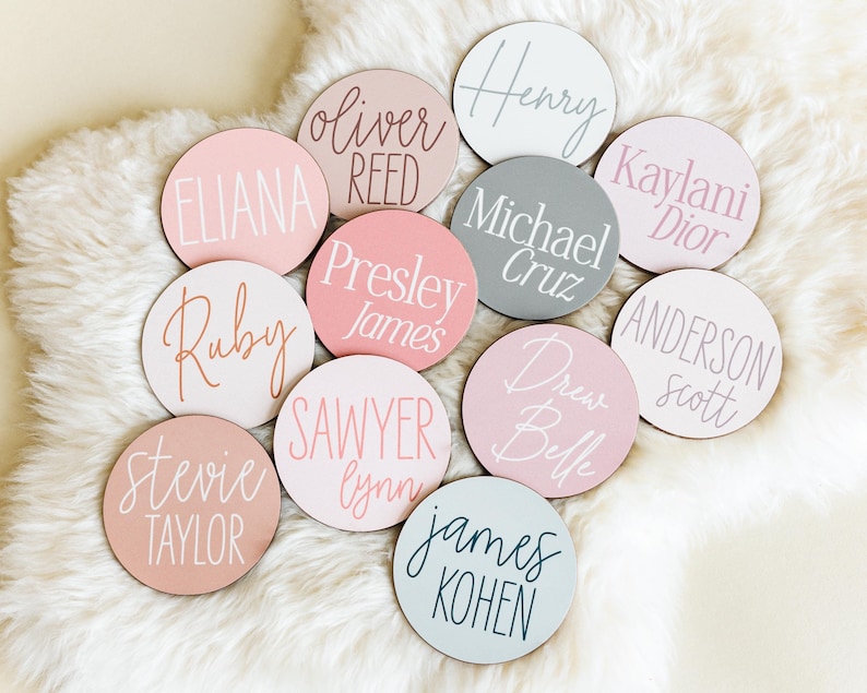 May include: A collection of 10 round wooden name tags with various names written in white, black, and pink calligraphy. The tags are on a white fluffy surface.