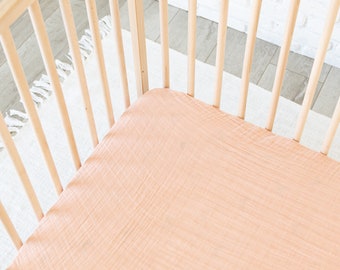 May include: A peach-colored fitted crib sheet made of soft, breathable fabric. The sheet is in a crib with wooden slats.
