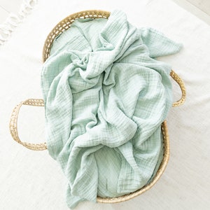 May include: A light green muslin swaddle blanket in a woven wicker basket with handles.