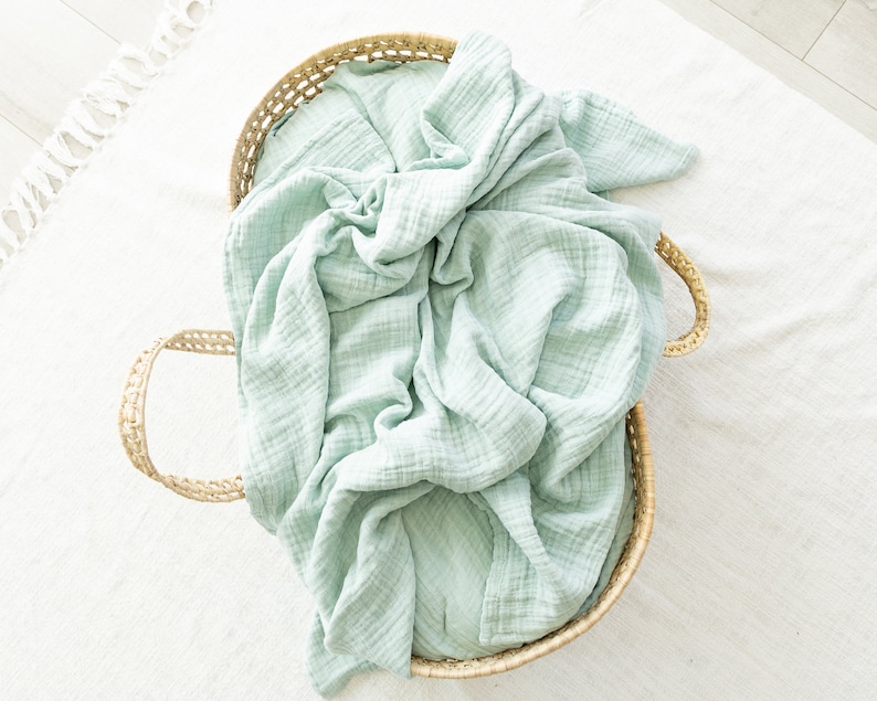 May include: A light green muslin swaddle blanket in a woven wicker basket with handles.