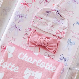 May include: A baby bedding set in pink with a bow pattern. The set includes a blanket, a knotted hat, and a headband. The blanket and hat are printed with the name "Charlotte Reese".