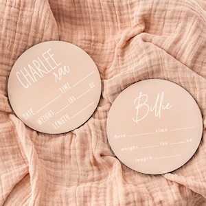 May include: Two round wooden plaques with pink backgrounds and white text. The top plaque says "CHARLEE Rae" and has spaces to fill in the date, weight in pounds and ounces, length in inches, and time of birth. The bottom plaque says "Billie" and has spaces to fill in the date, weight in pounds and ounces, length in inches, and time of birth.