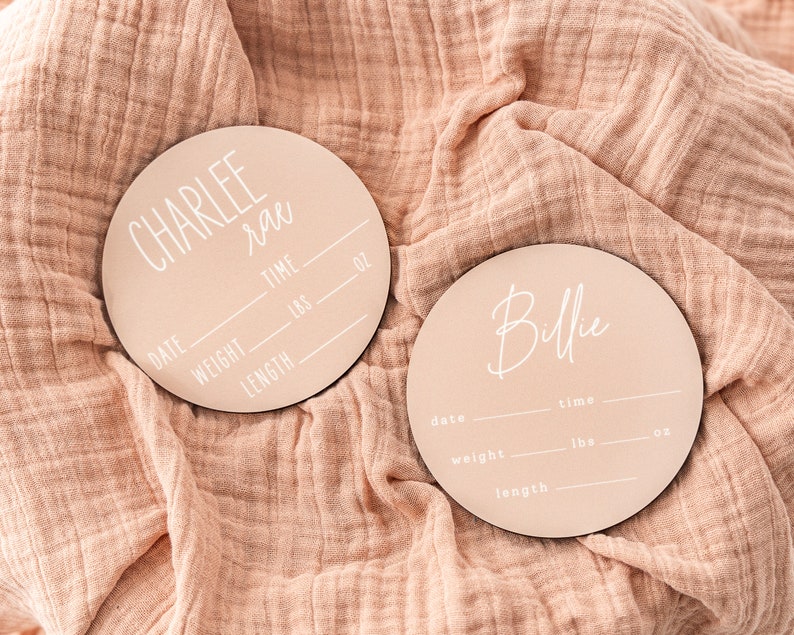 May include: Two round wooden plaques with pink backgrounds and white text. The top plaque says "CHARLEE Rae" and has spaces to fill in the date, weight in pounds and ounces, length in inches, and time of birth. The bottom plaque says "Billie" and has spaces to fill in the date, weight in pounds and ounces, length in inches, and time of birth.