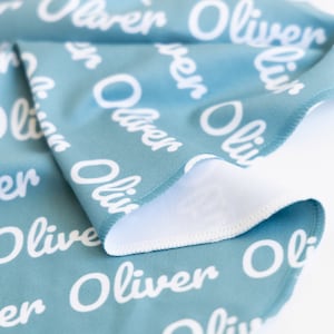 May include: A folded teal blanket with the name "Oliver" repeated in white script. The blanket has a white underside and a stitched edge. The fabric is soft and appears to be a baby blanket or similar item.
