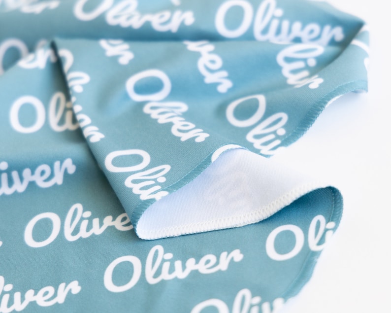 May include: A folded teal blanket with the name "Oliver" repeated in white script. The blanket has a white underside and a stitched edge. The fabric is soft and appears to be a baby blanket or similar item.