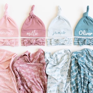 May include: Four baby sets in pink, mauve, light blue, and teal. Each set includes a knotted hat, a bow headband, and a swaddling blanket. The hats and headbands are personalized with names like "Summer," "Millie," "James," and "Oliver."