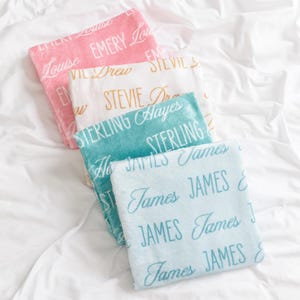 Op de afbeelding: Vier zachte, gepersonaliseerde dekens in roze, wit, blauwgroen en lichtblauw, elk met een andere naam in een sierlijk lettertype. De dekens zijn gerangschikt op een wit sprei, waardoor een gezellig en persoonlijk cadeau ontstaat.