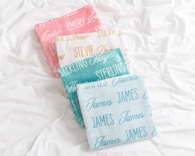 Op de afbeelding: Vier zachte, gepersonaliseerde dekens in roze, wit, blauwgroen en lichtblauw, elk met een andere naam in een sierlijk lettertype. De dekens zijn gerangschikt op een wit sprei, waardoor een gezellig en persoonlijk cadeau ontstaat.