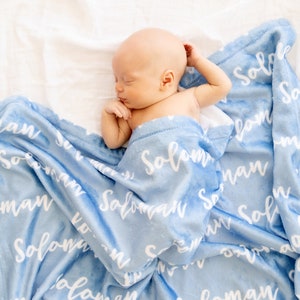 May include: A newborn baby is nestled in a soft, light blue blanket. The blanket is printed with the name "Solomon" in a white, cursive font. The baby is lying on a white surface, creating a serene and comforting scene.