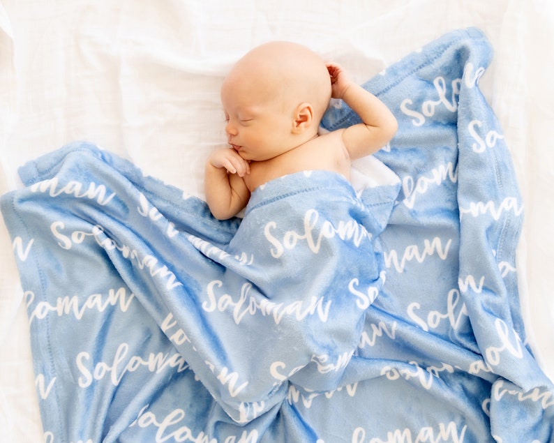 May include: A newborn baby is nestled in a soft, light blue blanket. The blanket is printed with the name "Solomon" in a white, cursive font. The baby is lying on a white surface, creating a serene and comforting scene.