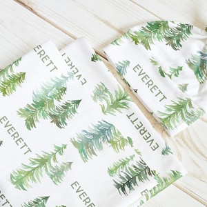 May include: A white baby blanket and hat set with a watercolor pine tree pattern and the name "EVERETT" printed on both items.