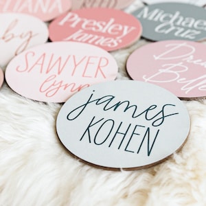 May include: A collection of round, wooden name tags with various names written in white and black calligraphy. The names include "Ruby", "Sawyer Lynn", "James Kohen", "Drew Belle", "Michael Cruz", and "Presley James".