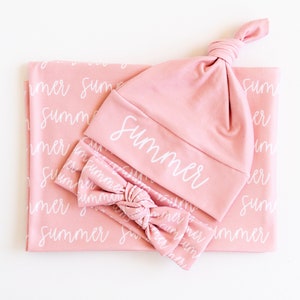 May include: A baby set in a soft pink color, including a blanket, a knotted hat, and a headband. The blanket and headband feature the word "summer" in a white script. The hat also has the word "summer" in white script.