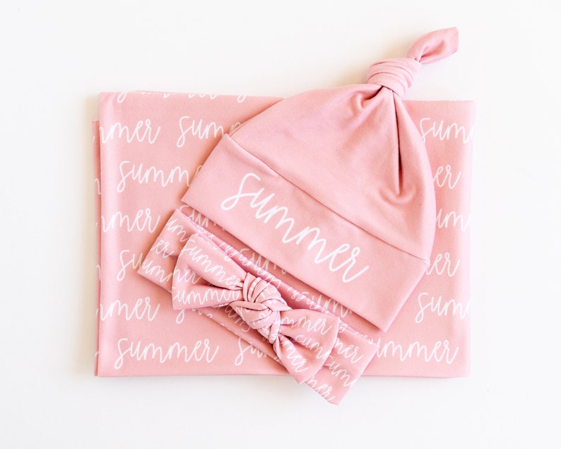 May include: A baby set in a soft pink color, including a blanket, a knotted hat, and a headband. The blanket and headband feature the word "summer" in a white script. The hat also has the word "summer" in white script.