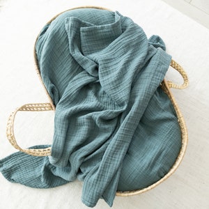 May include: A teal blue muslin swaddle blanket is folded and placed inside a woven wicker basket with handles.