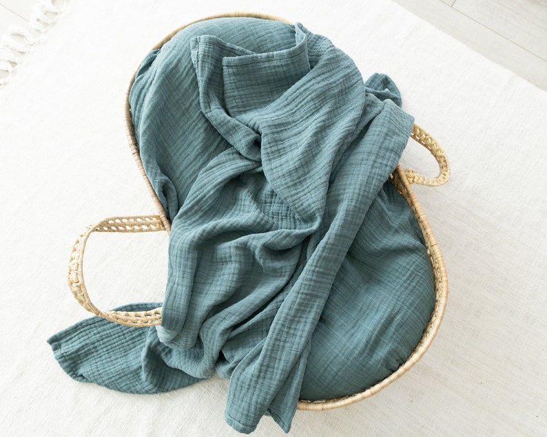 May include: A teal blue muslin swaddle blanket is folded and placed inside a woven wicker basket with handles.