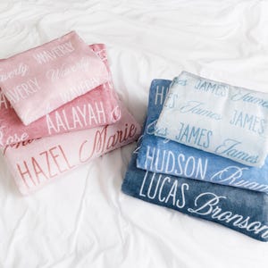 Op de afbeelding: Een stapel gepersonaliseerde dekens in roze en blauwe tinten. De roze dekens tonen namen als "Waverly" en "Hazel", terwijl de blauwe dekens namen als "James" en "Hudson" tonen. De namen zijn in een sierlijk lettertype.