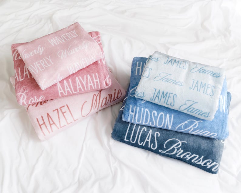 Op de afbeelding: Een stapel gepersonaliseerde dekens in roze en blauwe tinten. De roze dekens tonen namen als "Waverly" en "Hazel", terwijl de blauwe dekens namen als "James" en "Hudson" tonen. De namen zijn in een sierlijk lettertype.