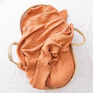 May include: A woven basket with a burnt orange muslin blanket inside. The basket has two handles on the sides.