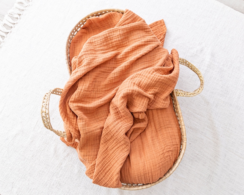 May include: A woven basket with a burnt orange muslin blanket inside. The basket has two handles on the sides.