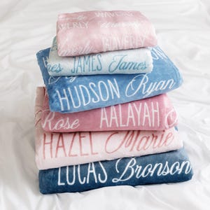May include: A stack of personalized blankets in various pastel colors, including pink, blue, and white. Each blanket is embroidered with a different name in a cursive font. The blankets are stacked on a white surface.
