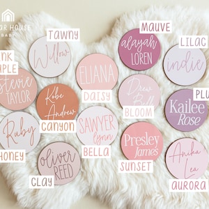 May include: A set of 16 round wooden name plaques with pink, white, and mauve paint. The plaques feature various names written in cursive script. The names include: Willow, Eliana, Alayah Loren, Indie, Drew Belle, Sawyer Lynn, Presley James, Anika Lea, Stevie Taylor, Kobe Andrew, Ruby Carson, Oliver Reed, Kailee Rose, Pink Maple, Daisy Bloom, and Honey Clay.