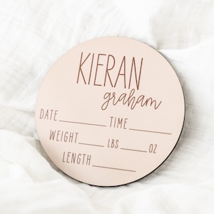 May include: A pink wooden baby announcement plaque with the name "Kieran graham" and spaces to fill in the date, time, weight in pounds and ounces, and length.