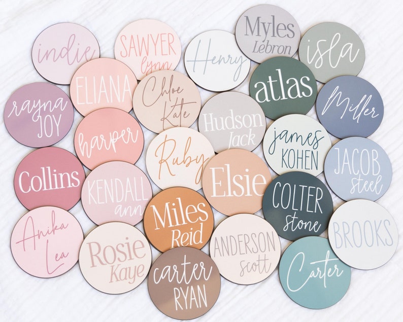 Op de afbeelding: Een verzameling van 20 ronde houten schijven met verschillende namen geschreven in wit schrift op een verscheidenheid aan pastelkleurige achtergronden. De namen zijn: Indie, Sawyer, Eliana, Harper, Collins, Kendall, Anika, Rosie, Miles, Carter, Henry, Chloe, Ruby, Elsie, Anderson, Hudson, James, Jacob, Isla en Carter.
