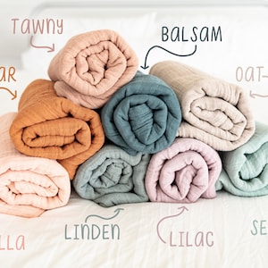 May include: A stack of eight rolled muslin swaddles in various colours, including cedar, tawny, balsam, oat, bella, linden, lilac, and seafoam.