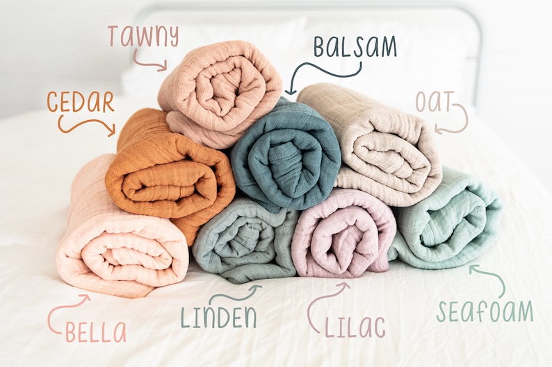 May include: A stack of eight rolled muslin swaddles in various colours, including cedar, tawny, balsam, oat, bella, linden, lilac, and seafoam.