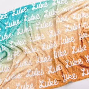 May include: A personalized beach towel with the name "Luke" repeated in white script across a gradient background. The colors transition from turquoise to orange, creating a warm, inviting aesthetic. The towel is soft and plush.