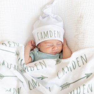 May include: A white baby blanket with green arrows and the name "Camden" repeated throughout. A baby wearing a white knotted beanie with the name "Camden" in green lettering is sleeping on the blanket.