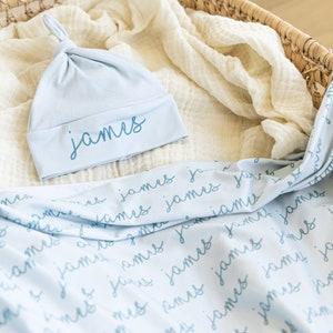 May include: A light blue baby hat and blanket set. The hat is knotted at the top and has the name "James" embroidered on the brim. The blanket is light blue with the name "James" repeated in a script font.
