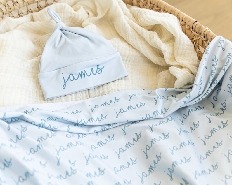 May include: A light blue baby hat and blanket set. The hat is knotted at the top and has the name "James" embroidered on the brim. The blanket is light blue with the name "James" repeated in a script font.