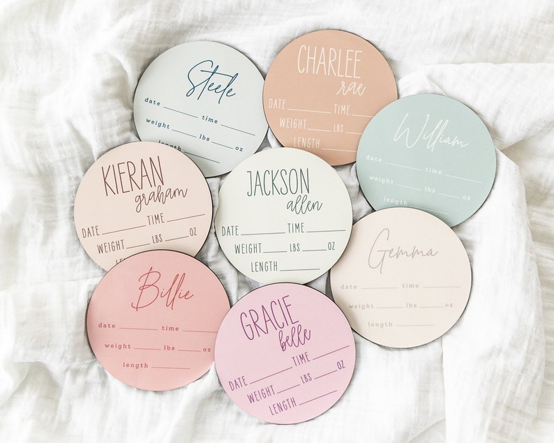May include: Seven round wooden plaques with pink, blue, and white backgrounds. Each plaque has a name written in cursive and includes spaces for date, time, weight in pounds and ounces, and length. The names are: Steele, Charlee Rae, William, Kieran Graham, Jackson Allen, Gemma, and Gracie Belle.