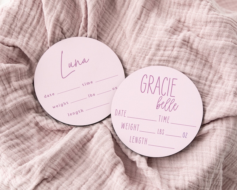 May include: Two pink round wooden baby announcement plaques with white text. One plaque says "Luna" and the other says "Gracie Belle". Both plaques have spaces to fill in the baby's date, time, weight in pounds and ounces, and length.