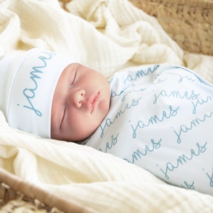 May include: A sleeping baby wrapped in a light blue swaddle blanket and matching hat. The name "James" is written in a script font on both the hat and blanket. The baby is nestled in a woven basket lined with a cream-colored fabric.