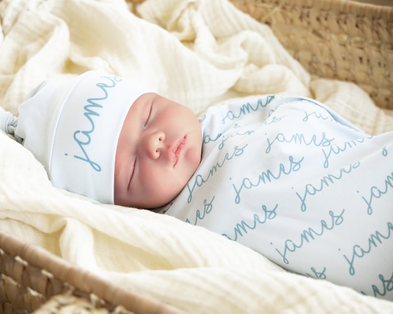 May include: A sleeping baby wrapped in a light blue swaddle blanket and matching hat. The name "James" is written in a script font on both the hat and blanket. The baby is nestled in a woven basket lined with a cream-colored fabric.