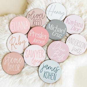 Op de afbeelding: Een verzameling van 12 ronde houten naamschildjes met verschillende namen in wit, roze en grijs schrift. De namen zijn Eliana, Oliver Reed, Presley James, Ruby, Sawyer Lynn, Stevie Taylor, Henry, Kaylani Dior, Michael Cruz, Drew Belle, Anderson Scott en James Kohen.